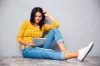 Mujer con jersey amarillo sentada en el suelo y leyendo ebook o tablet