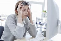 Mujer con jersey gris y camisa se sube las gafas para frotarse un ojo frente a un ordenador